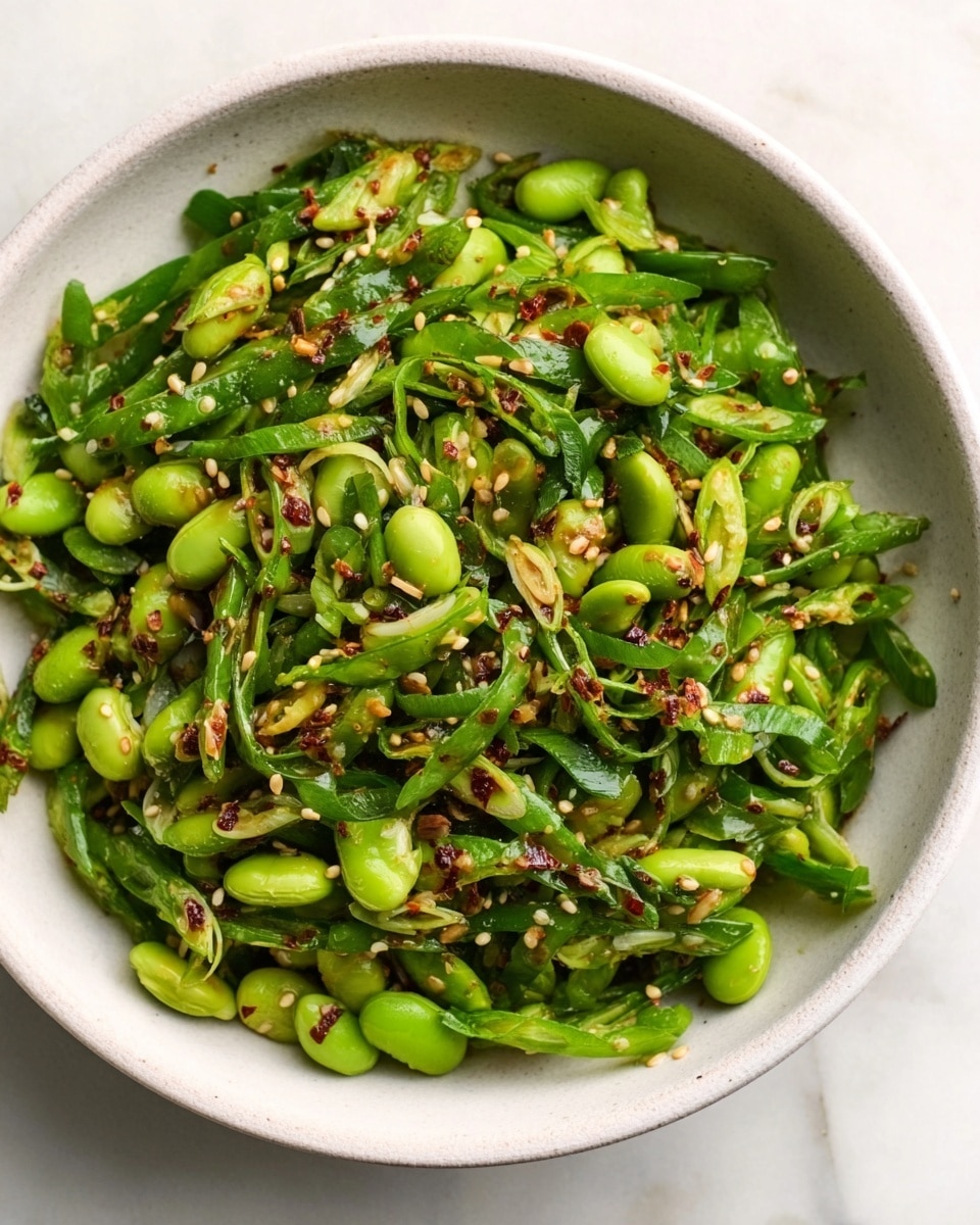 A close-up view of a fresh salad in a white bowl, mainly made of bright green edamame beans and thinly sliced green onions mixed together. The salad is sprinkled with white sesame seeds and small bits of red chili flakes, giving a textured look with a mix of smooth beans and crisp onion slices. There are small drops of dark soy-like sauce scattered across the salad, adding a glistening effect on the green ingredients. The bowl is placed on a white marbled surface, with some small pieces of salad and sauce visible near the edge. photo taken with an iphone --ar 4:5 --v 7