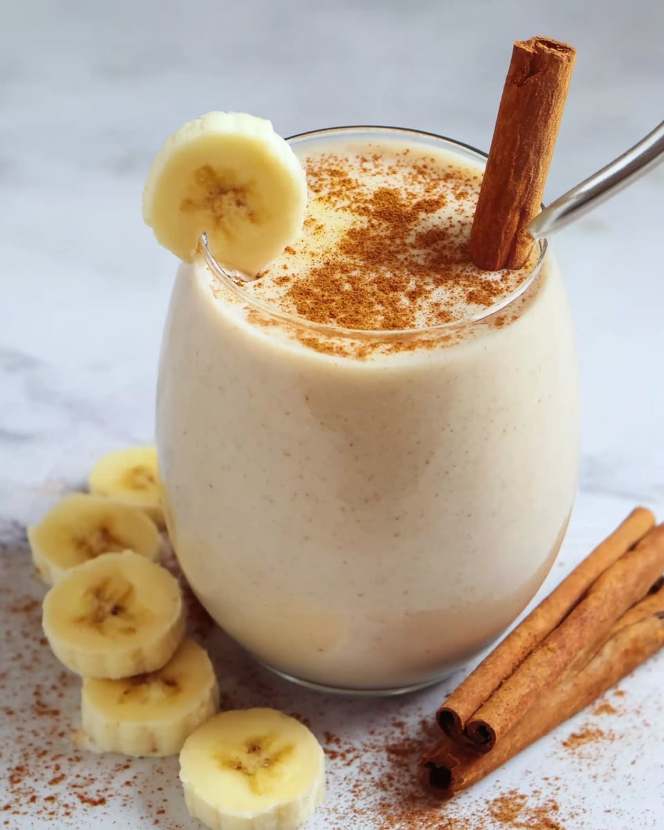 A tall clear glass is filled nearly to the top with a creamy pale beige banana smoothie that has a frothy texture. On the smoothie’s surface, two thin banana slices are lightly sprinkled with fine brown cinnamon powder. A stick of cinnamon stands upright inside the glass on the right side, while a single banana slice is placed on the rim on the left side. Around the base of the glass, several banana slices and two whole cinnamon sticks rest on a wooden surface with a warm tone. photo taken with an iphone --ar 4:5 --v 7