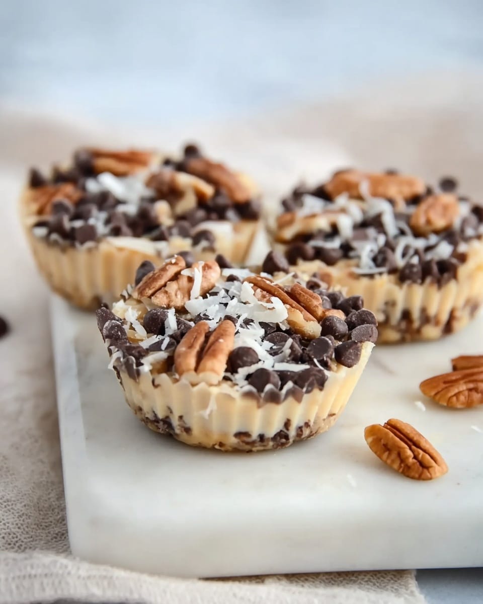 The image shows three small round treats placed on a white marble cutting board. Each treat has a creamy beige base layer, which is topped with a generous layer of dark brown chocolate chips scattered evenly. On top of the chocolate chips, there are pieces of light brown pecans and thin white shreds of coconut, creating a textured and colorful surface. The treats have a crinkled paper edge hinting they are baked or set in paper liners, and the background is softly blurred with a white and light tone, making the treats stand out clearly. photo taken with an iphone --ar 4:5 --v 7