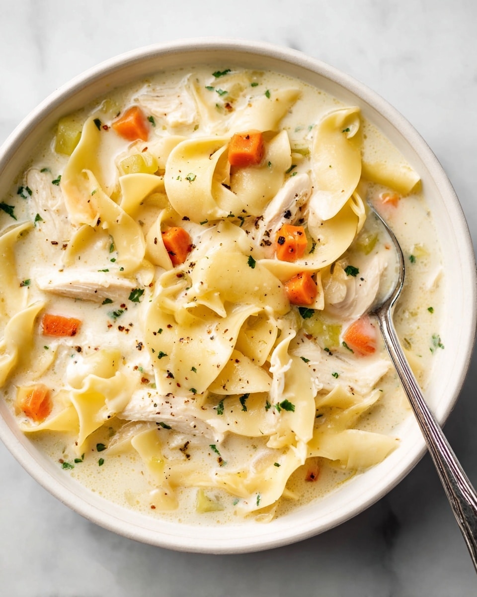 A bowl filled with creamy chicken noodle soup showing several layers: the top layer is smooth and thick cream broth, light beige in color; within this base layer are irregular wide egg noodles, pale yellow and soft-looking, spread across the bowl; scattered carrot cubes in bright orange and small celery bits in pale green add pops of color; chunks of white chicken meat are nestled among the noodles; fresh green parsley flecks are sprinkled on top; black pepper dots lightly enhance the surface; a silver spoon rests inside the bowl, dipping into the soup. The bowl is white and sits on a white marbled surface. photo taken with an iphone --ar 4:5 --v 7