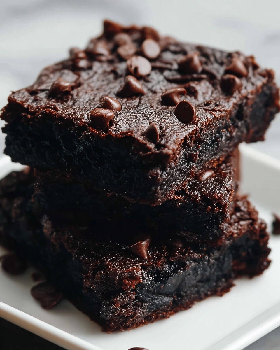The image shows two dark brown chocolate brownies stacked on top of each other on a white plate, set on a white marbled surface. Each brownie has a shiny, cracked top layer sprinkled with small, glossy dark chocolate chips scattered evenly. The bottom layer of each brownie appears dense and moist with a slightly rough texture. The top brownie is positioned at a slight angle over the bottom brownie, creating a clear view of the thick layers and rich chocolate texture. Photo taken with an iphone --ar 4:5 --v 7