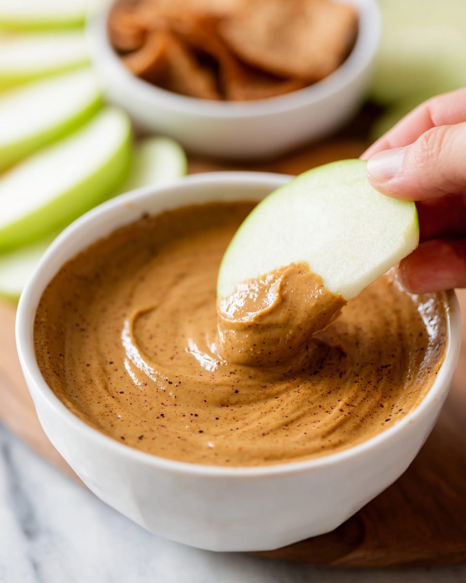 A white bowl sits filled with a smooth, light brown creamy dip sprinkled evenly with a darker brown powder on top, creating a slightly textured surface with gentle swirls in the center. Behind the bowl, there are several slices of bright green apple arranged in overlapping layers on a white marbled surface. To the right side, a white plate holds light brown square-shaped crackers stacked loosely. A folded white cloth is placed near the bowl, completing the simple and fresh arrangement. Photo taken with an iphone --ar 4:5 --v 7