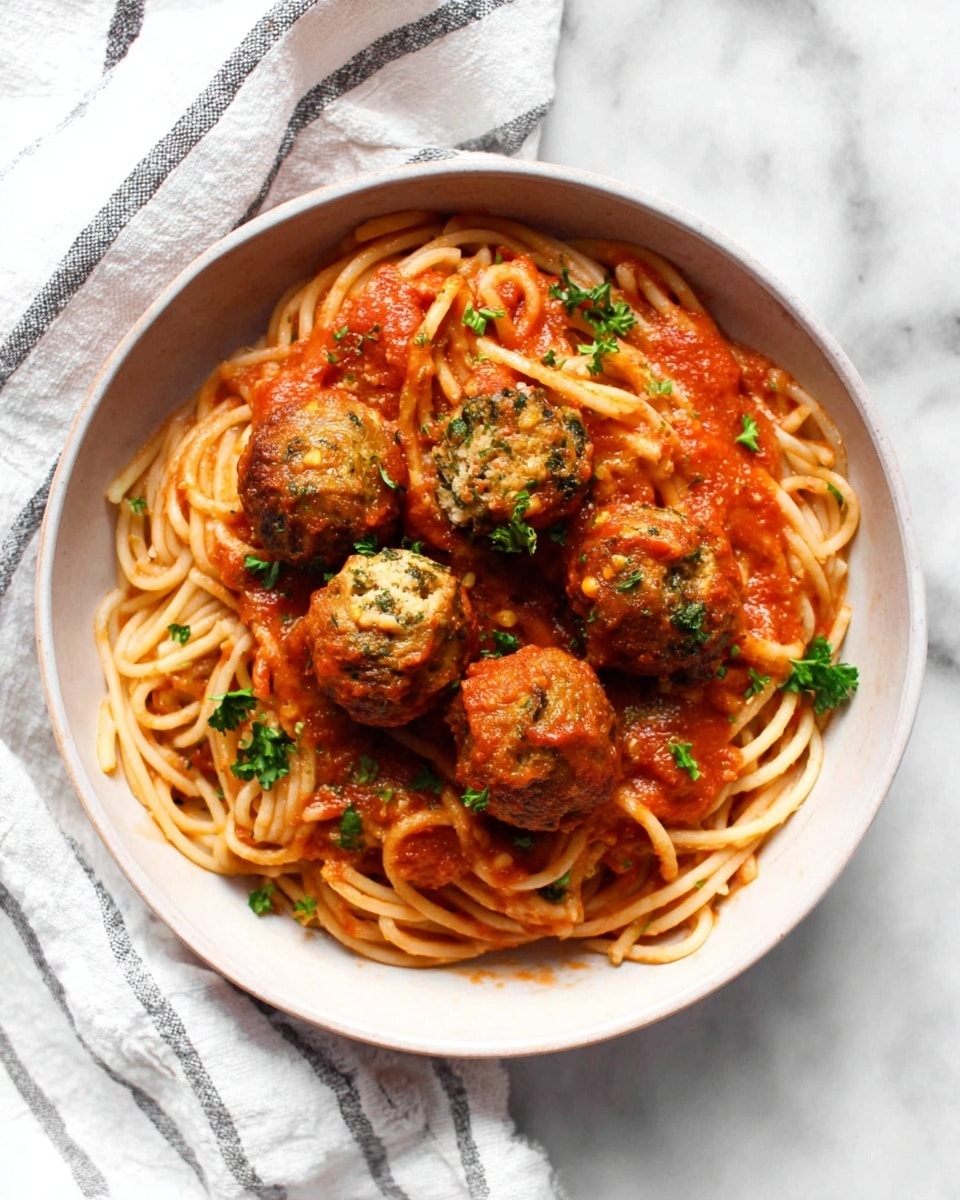 A white bowl is filled with a base layer of cooked spaghetti noodles that are coated lightly with red tomato sauce. On top, five golden-brown meatballs with hints of green herbs inside are placed in the center. The meatballs are partially covered with more thick red sauce, and fresh green parsley leaves are sprinkled over everything, adding contrast. The bowl sits on a white marbled surface with a white cloth featuring gray stripes underneath one side. Photo taken with an iphone --ar 4:5 --v 7