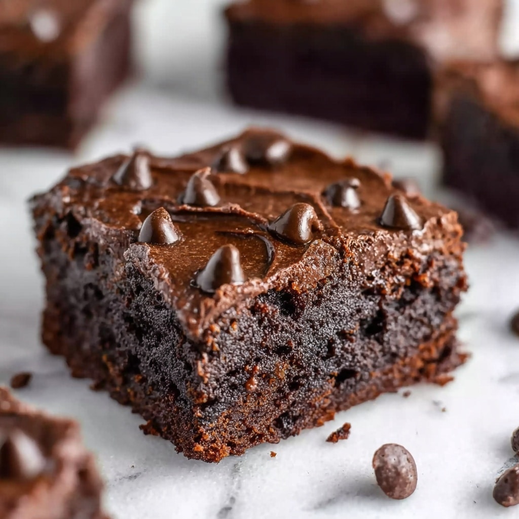 A close-up of a single square piece of chocolate brownie on a white marbled surface, showing two layers: a dense, dark brown bottom layer with a moist, cake-like texture, and a thick, darker top layer of glossy chocolate frosting dotted with shiny chocolate chips. The edges of the brownie are slightly rough, and in the background, there are blurred, smaller pieces of similar brownies scattered. photo taken with an iphone --ar 4:5 --v 7