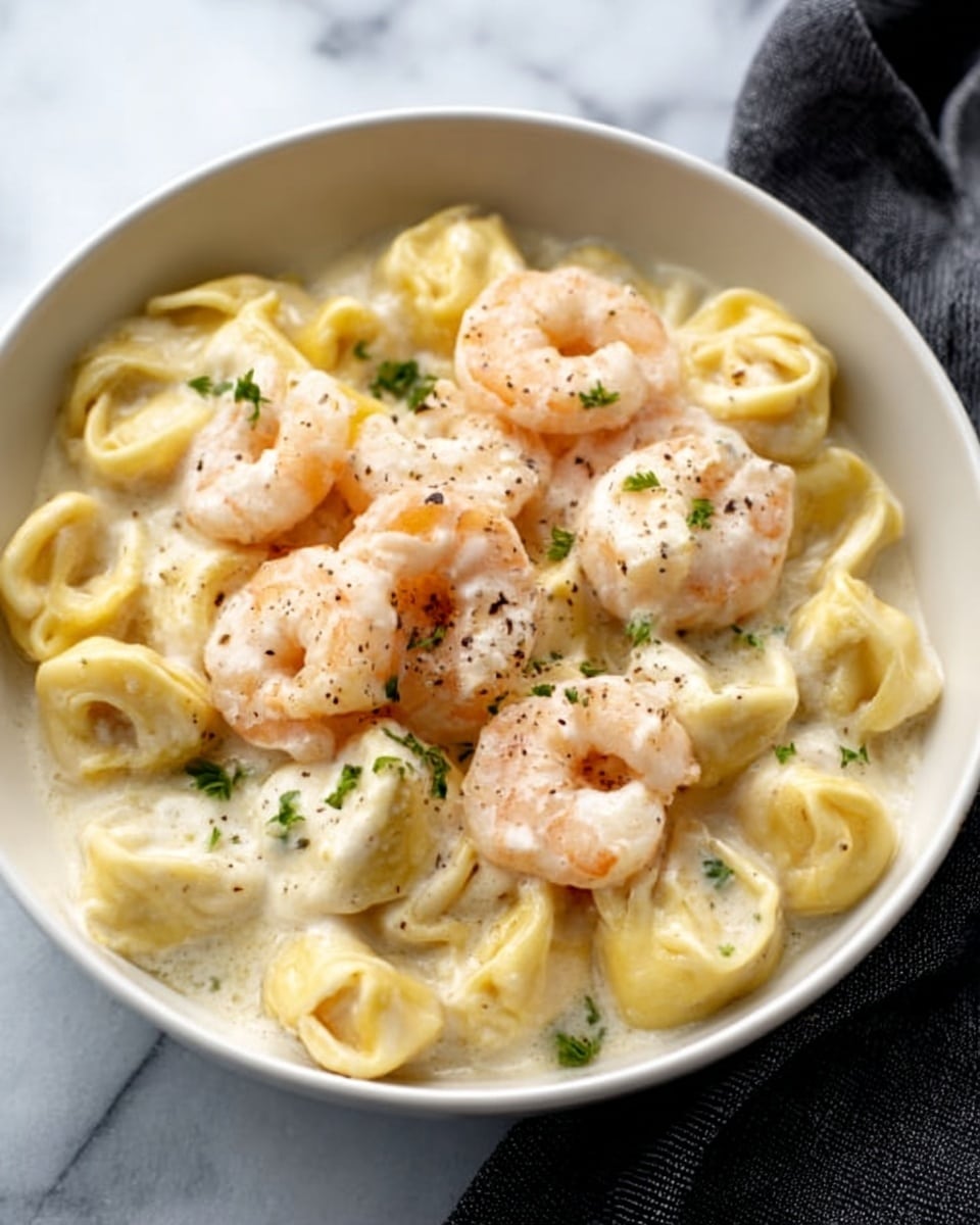 This image shows a close-up of a creamy shrimp and tortellini dish in a black pan. The base layer is a thick white sauce with a smooth texture, covering the entire pan. Scattered on top are light pink cooked shrimp, each with a slightly curved shape and a shiny surface. Interspersed among the shrimp are pale yellow tortellini pieces, their folded, round shapes visible, lightly coated in the sauce. Small bits of finely chopped green herbs are sprinkled evenly across the dish, adding color contrast. The dish looks rich and fresh. Photo taken with an iphone --ar 4:5 --v 7