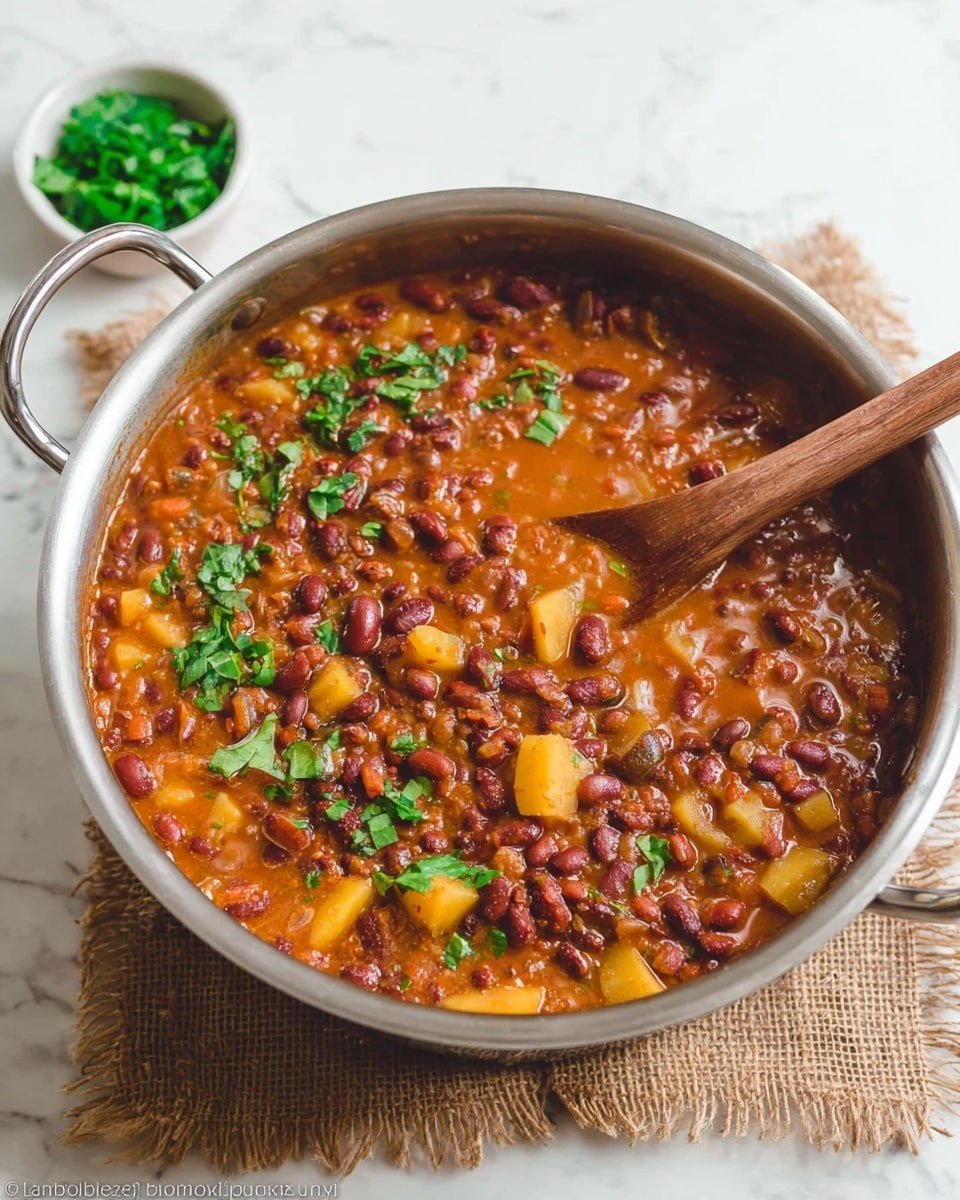 A metal pot filled with a thick stew showing several layers: the bottom layer is a rich, orange-brown broth with a slightly oily texture; scattered throughout are many small dark red beans and larger beige beans, creating a chunky and hearty look; among the beans are soft yellowish potato pieces adding texture contrast; bright green leaf pieces are sprinkled on top, giving a fresh touch; a wooden spoon is partially submerged in the stew, angled towards the right side of the pot; the pot rests on a rough burlap cloth over a white marbled surface; a small white bowl with more green leaves is in the blurred background at the top left. photo taken with an iphone --ar 4:5 --v 7