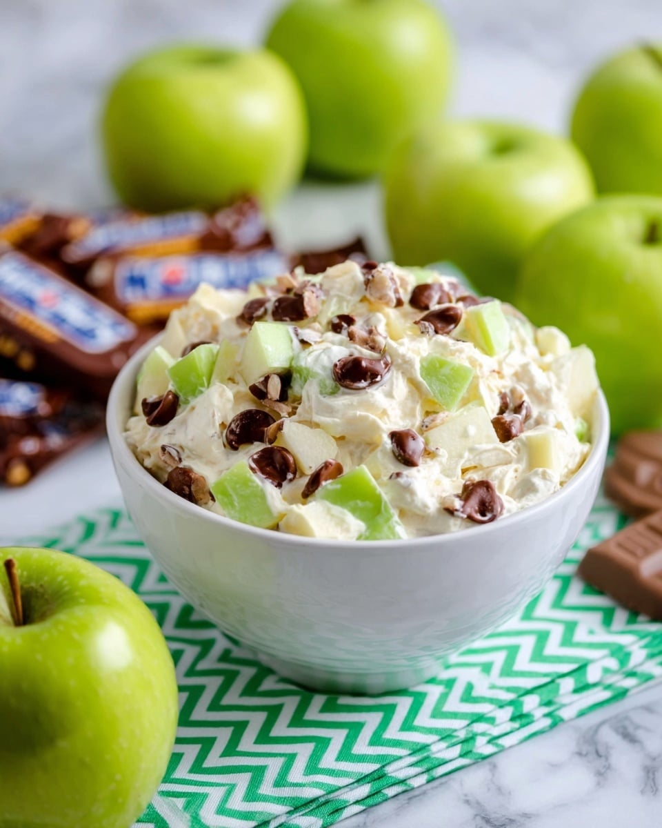 There is a close-up of a spoon with a mix of creamy white whipped topping, small cubes of green apple, white marshmallows, and chunks of chocolate. The spoon holds this mix above a white bowl that is full of the same ingredients. The topping looks smooth and soft, with a light drizzle of caramel or similar sauce on top. The background shows a white marbled surface blurred out. photo taken with an iphone --ar 4:5 --v 7