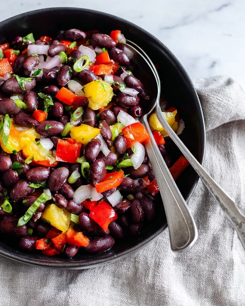 A close-up of a bowl filled with a colorful salad consisting of three main layers: a base layer of dark brown beans, scattered with bright red chopped bell peppers as the second layer, and topped with yellow pineapple chunks mixed with green sliced scallions. Two silver spoons rest partially inside the white bowl, which sits on a white marbled surface next to a light beige cloth. The salad looks fresh and vibrant with many small pieces visible. photo taken with an iphone --ar 4:5 --v 7