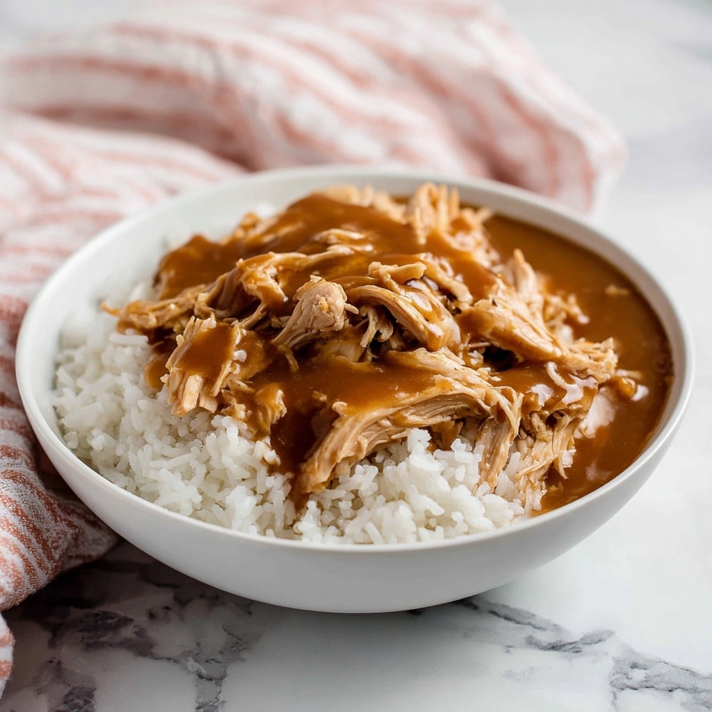 A white bowl filled with a base layer of white rice, topped with shredded pieces of cooked chicken that are covered with thick brown gravy. The chicken looks tender and moist, with the gravy smoothly coating the meat in a glossy texture. The bowl is placed on a white marbled surface with a faint pink and white striped cloth in the background. photo taken with an iphone --ar 4:5 --v 7