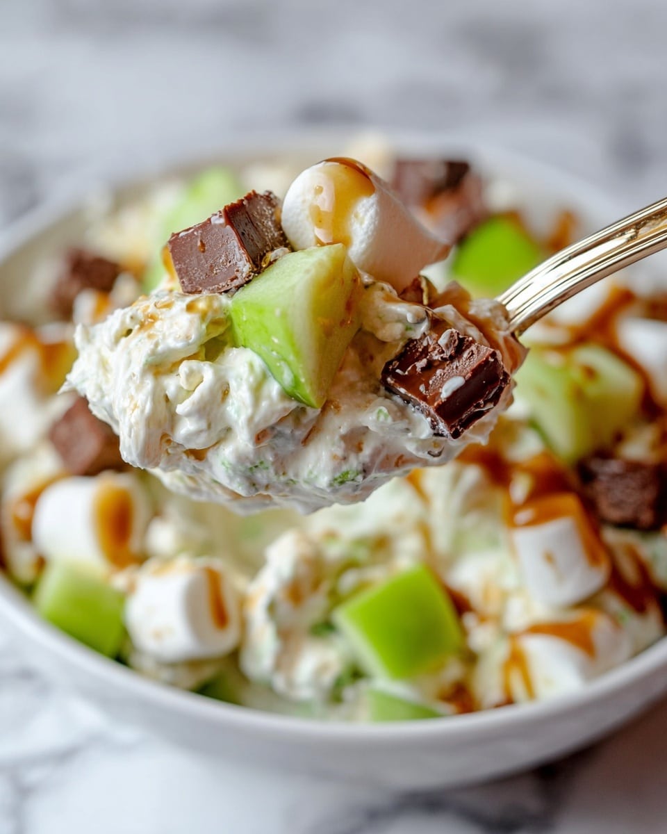 A white bowl filled with a creamy mixture that has visible layers of white cream, light beige chunks, and bright green apple pieces scattered throughout, topped with small, dark brown chocolate-covered nut candy pieces as a final layer. The bowl sits on a green and white zigzag cloth with a single green apple in front, surrounded by more bright green apples and some chocolate candy bars in the background, all placed on a white marbled texture surface. Photo taken with an iphone --ar 4:5 --v 7