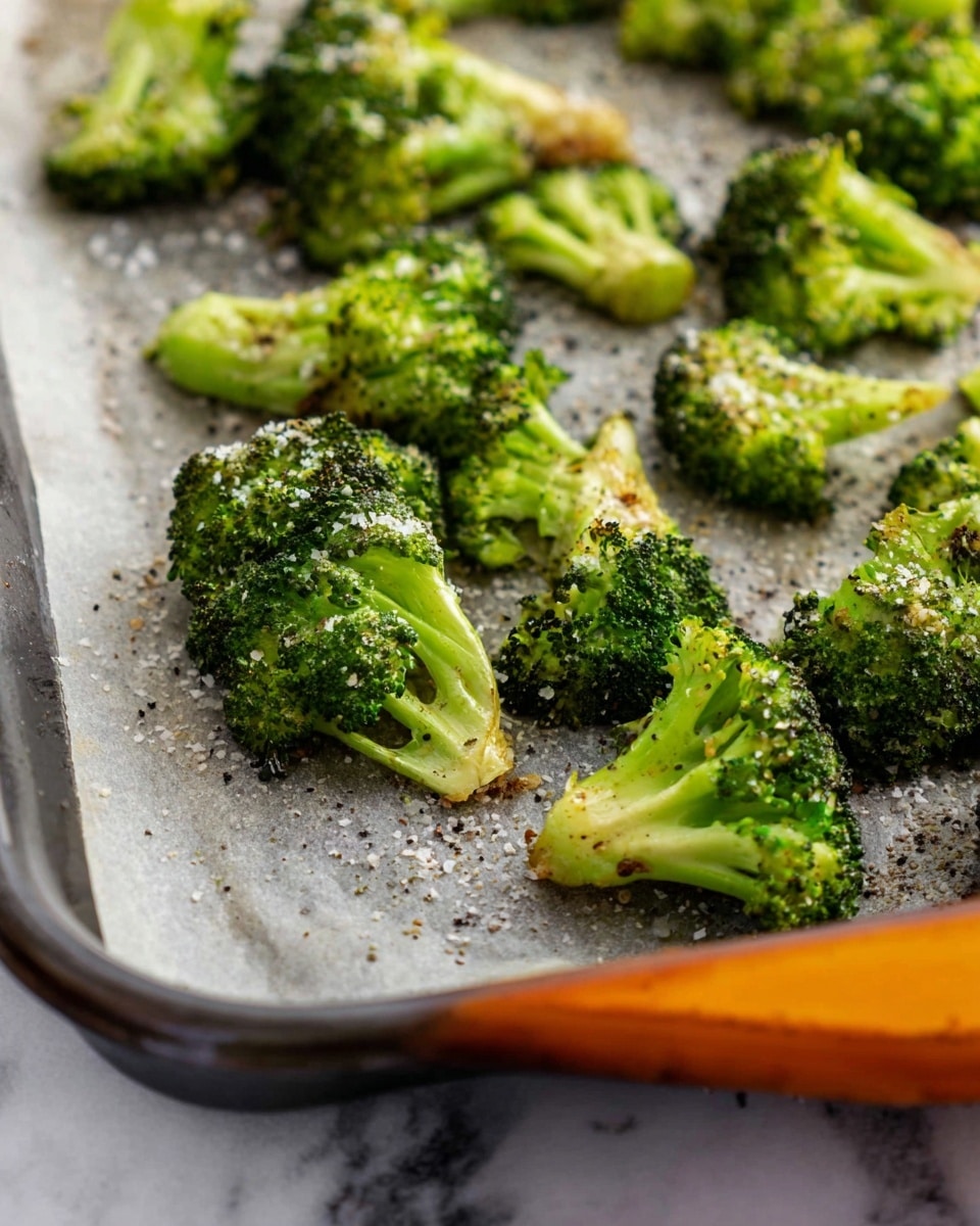 The image shows small bright green broccoli florets spread out on a baking tray lined with parchment paper. The florets have a slightly roasted texture with hints of crunchy edges and are sprinkled unevenly with coarse white salt and black pepper. The baking tray has a gray edge with an orange silicone handle visible in the corner, and everything is placed on a white marbled texture surface. The focus is mainly on the broccoli in the foreground, showing its fresh and cooked details clearly. photo taken with an iphone --ar 4:5 --v 7