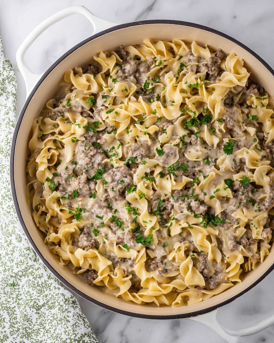 The image shows a large white pan filled with a creamy pasta dish made of three layers. The bottom and middle layers are soft, twisted egg noodles in pale yellow. Between and on top of the noodles, there is a thick sauce mixed with small, cooked ground beef pieces in brown and grayish tones. The creamy sauce is light beige and coats the noodles and beef evenly, giving a rich, smooth texture. Small bits of fresh green herbs are sprinkled on top, adding a pop of color over the whole dish. The pan is placed on a white marbled surface, and a part of a white and green patterned cloth is visible in the corner. Photo taken with an iphone --ar 4:5 --v 7