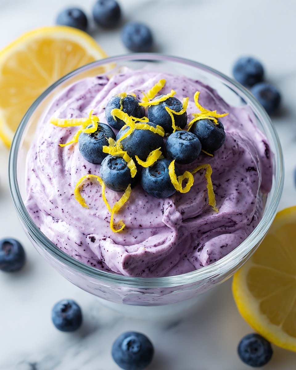 A clear glass bowl filled with a thick, creamy, light purple mousse that has tiny dark purple specks throughout. On top, there are about eight fresh, plump blueberries arranged in the center along with thin, bright yellow lemon zest curls scattered evenly. The bowl sits on a white marbled surface with a few blueberries and a half lemon nearby, adding fresh, natural elements to the scene. photo taken with an iphone --ar 4:5 --v 7