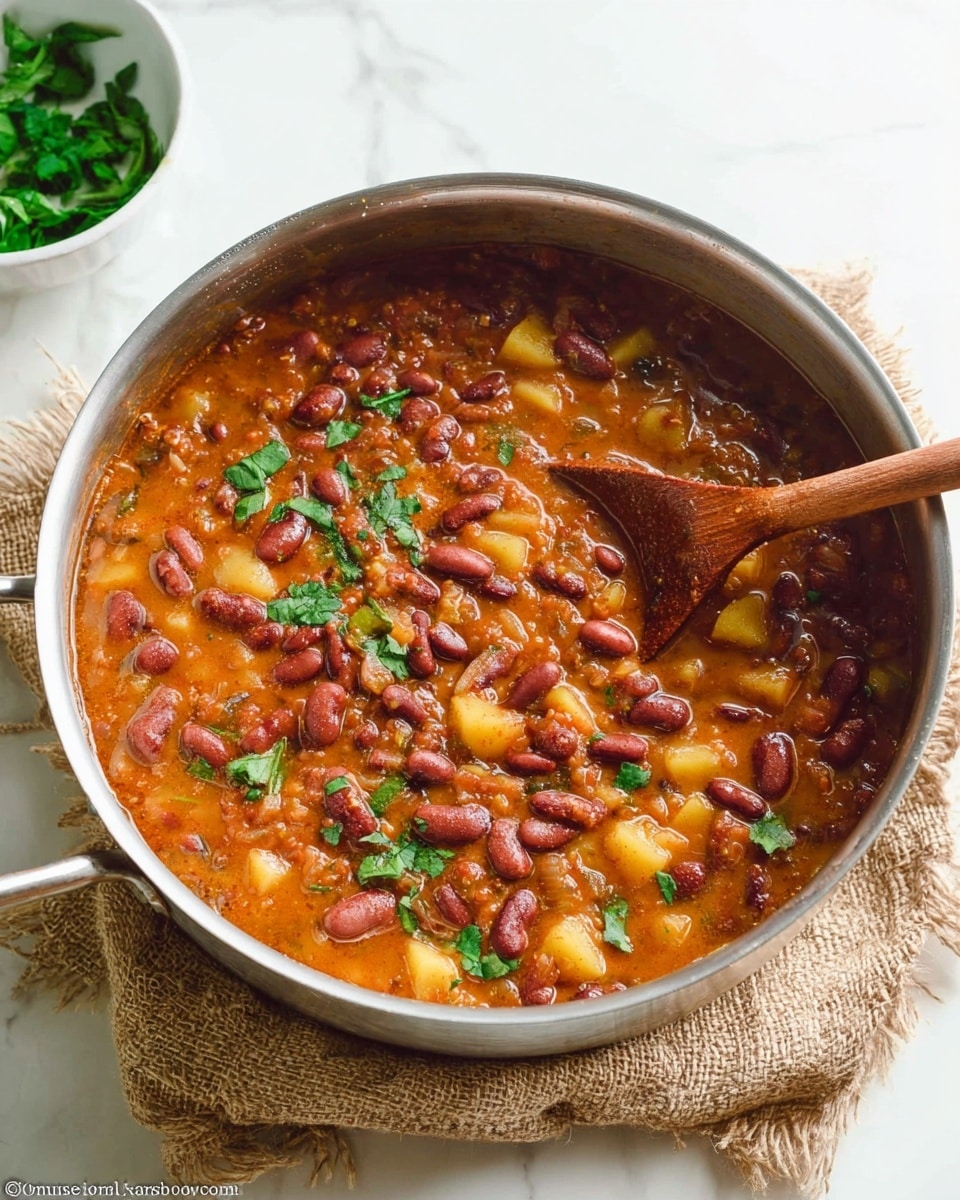 The image shows a white bowl filled with thick red bean stew. The stew has a rich, reddish-brown color with visible whole red beans and small chunks of cooked vegetables, giving it a chunky texture. On top of the stew, there is a small pile of fresh green cilantro leaves adding a splash of bright green color. A silver spoon is placed inside the bowl, partially submerged in the stew. The bowl is set on a white marbled surface. photo taken with an iphone --ar 4:5 --v 7