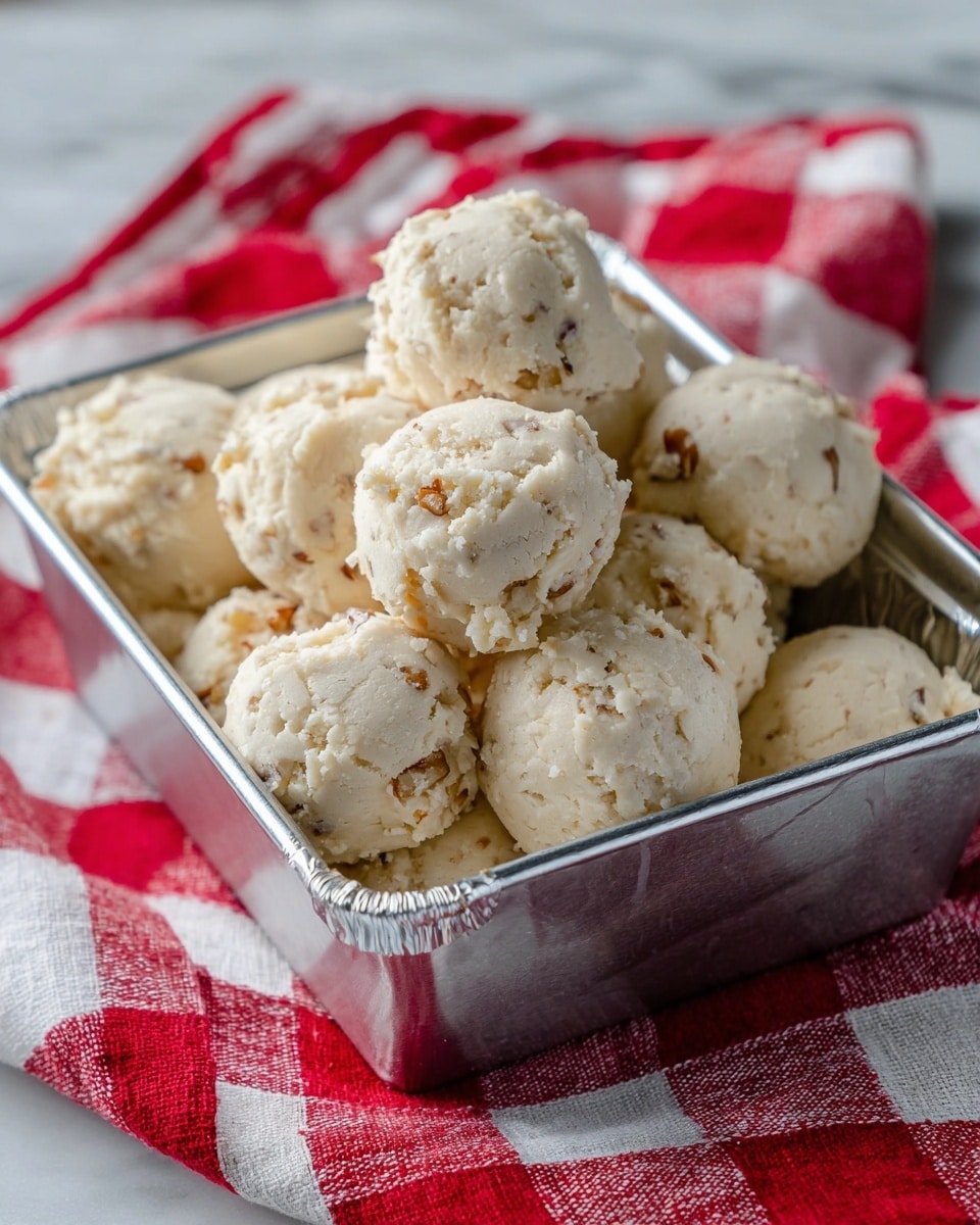 The image shows two trays filled with evenly spaced, round scoops of a creamy, light beige dough that has a crumbly texture on the edges. The top tray is a shiny silver baking pan holding fifteen dough scoops arranged in neat rows, with each scoop having a rough, uneven surface and bits of dough spread slightly around the base. Below it is a silver baking sheet lined with white parchment paper, holding six similar dough scoops. The background is a white marbled texture surface beneath the trays. photo taken with an iphone --ar 4:5 --v 7