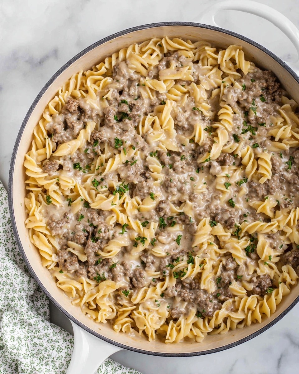A top-down view of a white pan filled with creamy beef and egg noodle pasta. The dish has two main layers: the base layer of pale yellow egg noodles with a soft wavy texture and the top layer of small browned ground beef pieces mixed evenly in a light creamy sauce. Small green parsley flakes are sprinkled on top for color contrast. A white spatula with a wooden handle is partly inside the pan on the right side, lifting some noodles and beef. The pan is set on a white marbled surface with a small salt shaker visible in the upper right corner and a green-patterned cloth partially seen on the bottom left. photo taken with an iphone --ar 4:5 --v 7