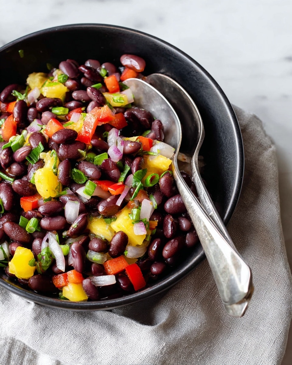 A close-up view of a black bowl filled with a colorful bean salad made primarily of small dark purple beans as the base layer. Mixed in are bright red chopped bell pepper pieces, small yellow fruit chunks, and slices of white onion scattered throughout. Fresh green herbs and scallions add small touches of color on top. Two silver spoons rest inside the bowl, with the handles leaning on a light gray linen cloth. The bowl is placed on a white marbled surface. Photo taken with an iphone --ar 4:5 --v 7