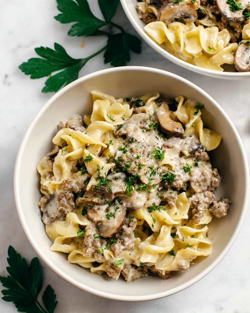 The image shows a close-up of two white bowls filled with a creamy pasta dish placed on a white marbled surface. The pasta has three visible layers: the first layer is curly egg noodles with a light yellow color and soft texture; the second layer is a creamy white sauce mixed with ground meat pieces and sliced brown mushrooms, giving a rich and thick look; the third layer is sprinkled fresh green parsley on top, adding a touch of color and freshness. One bowl is fully visible in the front center, while another bowl is partially visible in the upper right corner with similar contents. Two green parsley leaves lie on the white marbled surface near the bowls. photo taken with an iphone --ar 4:5 --v 7