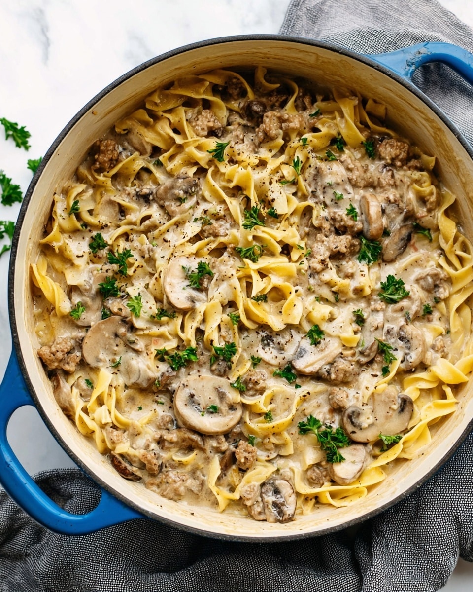 A large white pot with a blue handle holds creamy pasta with several layers of soft, yellow egg noodles mixed with pieces of browned ground meat and sliced brown mushrooms, all coated in a thick, light beige sauce. Small green parsley leaves are sprinkled throughout the dish, with a few placed on top for garnish, adding a fresh pop of color. The mixture has a slightly glossy texture, showing the richness of the sauce, and some black pepper is visible on the surface. The pot sits on a white marbled texture with a folded gray cloth beside it. Photo taken with an iphone --ar 4:5 --v 7