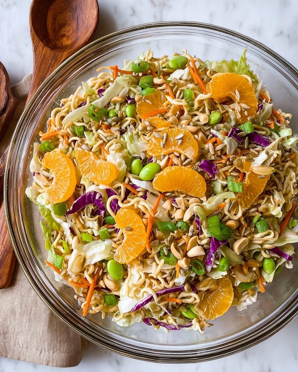 A close-up view of a white bowl filled with a colorful noodle salad sitting on a white marbled surface. The dish has several layers and textures: the bottom is a mixture of green edamame beans, shredded white and purple cabbage, and thin orange carrot strips. Scattered on top are light tan, curly dried ramen noodles adding a crunchy texture. Bright orange mandarin segments are spread throughout, adding a fresh and juicy contrast. The whole salad looks vibrant and fresh, with some loose noodles and beans scattered around the bowl. In the blurry background, green onions and wooden kitchen utensils are visible. Photo taken with an iphone --ar 4:5 --v 7