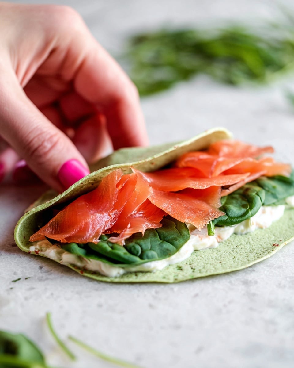 A close-up image shows a green spinach wrap being folded by a woman's hand with pink nail polish. The wrap has three visible layers: a base layer of creamy white spread, a middle layer of fresh dark green spinach leaves, and a top layer of thin, bright orange-red slices of smoked salmon spread unevenly. The wrap is on a white marbled surface, with some scattered green herbs blurred in the background. The photo taken with an iphone --ar 4:5 --v 7