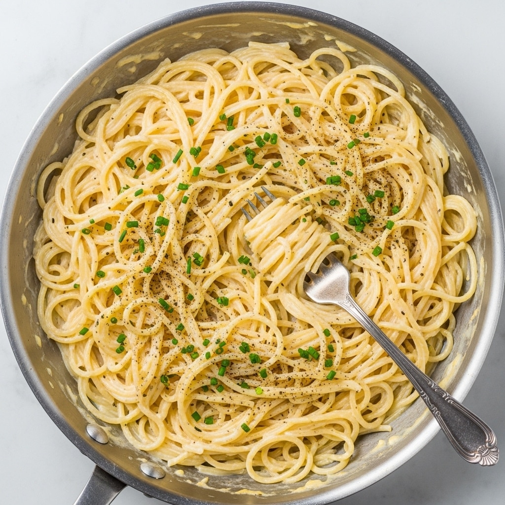 The image shows a pan filled with creamy spaghetti coated in a white sauce, with a slight yellow tint. The noodles are long and intertwined, spread evenly across the pan. Small green chive pieces are sprinkled on top, and there is a light dusting of black pepper adding texture and contrast. A silver fork with an ornate handle is placed inside the pan, twirling some spaghetti around its tines. The pan rests on a white marbled surface. Photo taken with an iphone --ar 4:5 --v 7
