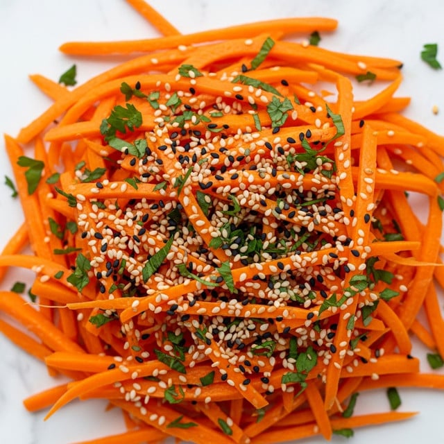 The image shows a close-up of a bright orange carrot salad made of thin, long carrot strips layered closely together, giving a fresh and crunchy texture. Scattered on top are small, evenly distributed white and black sesame seeds adding contrast and texture, while finely chopped green herbs are strewn across the salad, providing a fresh, leafy green splash. The background is a white marbled texture. photo taken with an iphone --ar 4:5 --v 7
