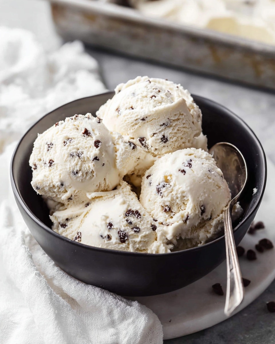 A close-up of a rectangular metal container filled with soft, creamy white ice cream mixed with small dark chocolate chips spread evenly throughout. The ice cream has a smooth but slightly textured surface with swirl marks. In the middle, a shiny metal ice cream scoop is digging into the ice cream, holding a rounded scoop filled with the mix of white cream and chocolate bits. The container rests on a surface with a white marbled texture. Photo taken with an iphone --ar 4:5 --v 7