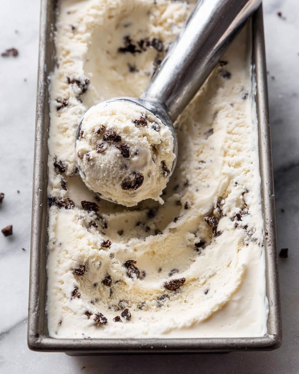 The image shows a black bowl filled with four scoops of creamy white ice cream with small dark chocolate chips scattered evenly throughout each scoop. The ice cream has a smooth, slightly fluffy texture with some soft peaks and creases. A silver spoon is placed inside the bowl on the right side, slightly buried in the ice cream. The bowl sits on a white marbled surface with a white cloth draped casually around it, while a blurry metal tray with more ice cream and scooping utensils is visible in the background. photo taken with an iphone --ar 4:5 --v 7