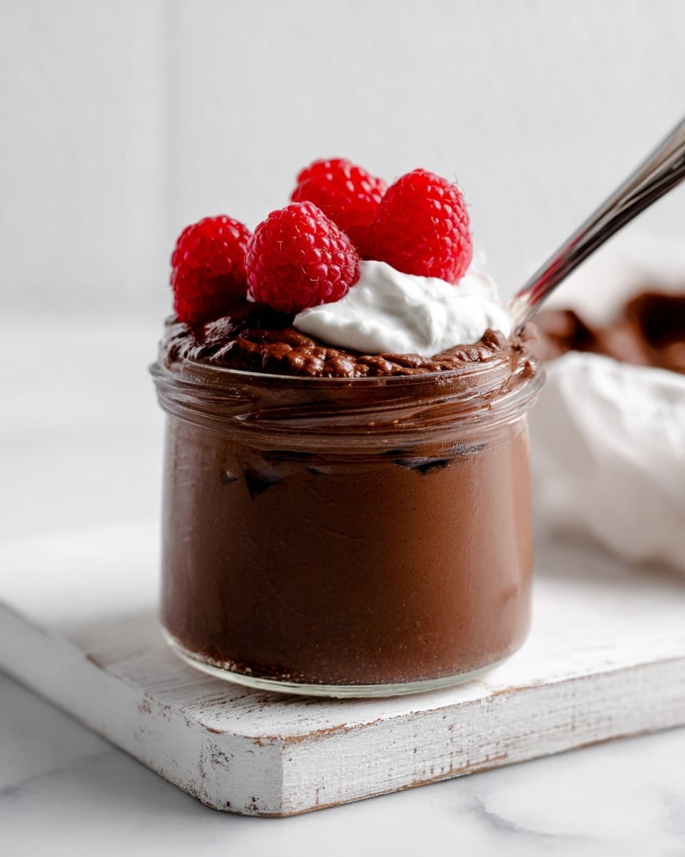 The image shows a small glass filled with three layers: a dark and smooth chocolate mousse at the bottom, a dollop of white whipped cream sitting on top to the right side, and three bright red raspberries placed on the cream and slightly on the mousse. The glass rests on a white marbled surface with a silver spoon on the bottom right holding some chocolate mousse. Other glasses with similar layers and toppings are partially visible around the main glass. Photo taken with an iphone --ar 4:5 --v 7