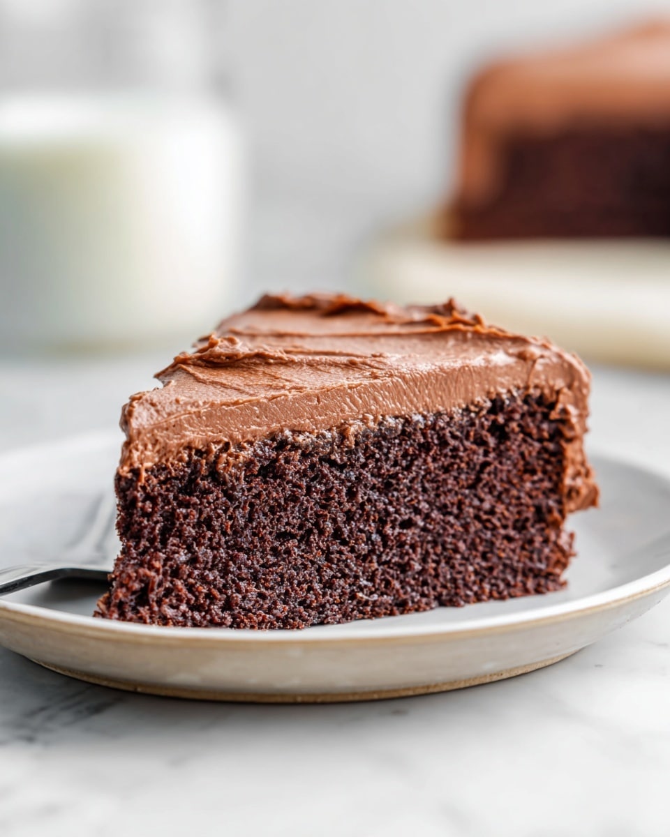 A single slice of chocolate cake is shown resting on a white plate with brown speckles. The cake has two visible layers: the bottom layer is a rich, dark brown chocolate sponge with a moist and slightly crumbly texture, while the top layer is a thick, smooth, and creamy chocolate frosting spread evenly with soft, curved swirls. The background is blurred and features a white marbled texture surface with a glass of milk softly out of focus. Photo taken with an iphone --ar 4:5 --v 7