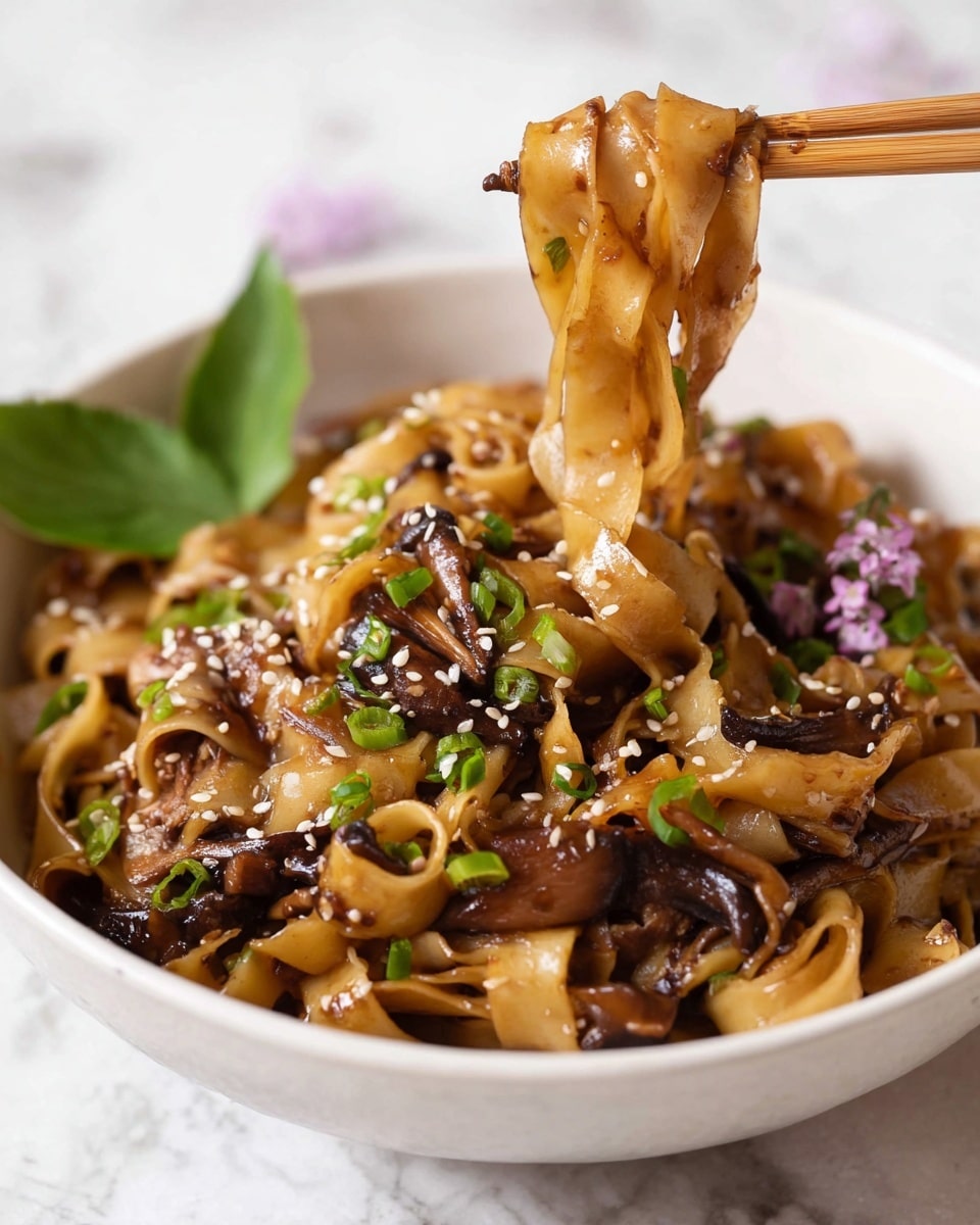 A bowl filled with wide, flat noodles mixed with dark brown cooked mushrooms and bits of green onions scattered on top. The noodles and mushrooms are coated with a glossy brown sauce, and white sesame seeds are sprinkled all over. A pair of chopsticks holds a small bundle of the noodles and mushrooms above the bowl. There is a small green leaf with sesame seeds and tiny purple flowers placed on the side inside the bowl. The bowl is white and sits on a white marbled surface. Photo taken with an iphone --ar 4:5 --v 7