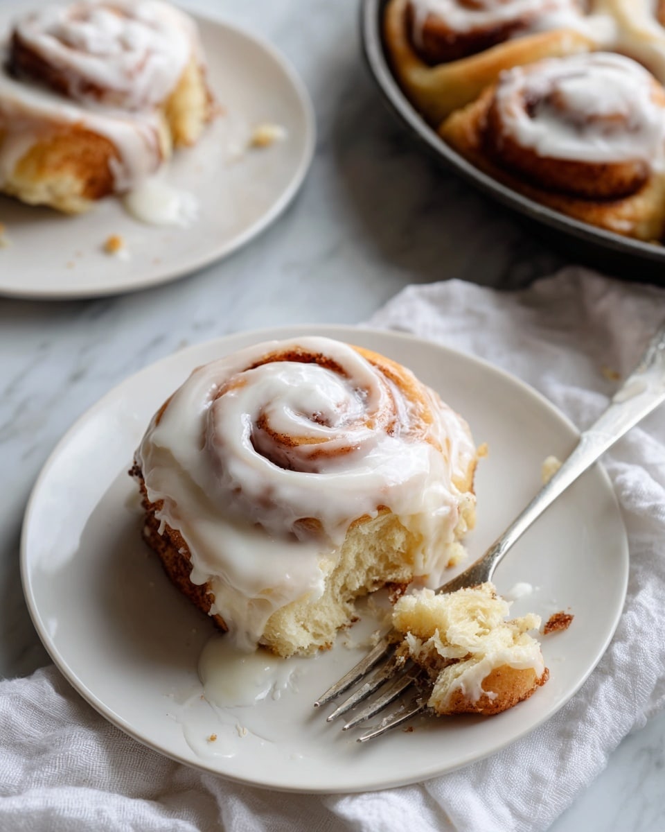A close-up of a white plate with a cinnamon roll covered in thick, creamy white icing, showing one layer of golden-brown dough spiraled with cinnamon and sugar. The cinnamon roll is partially eaten, with a silver fork placed on the right side of the plate. In the background, there is a black pan filled with more cinnamon rolls glazed with the same white icing. Another white plate with a cinnamon roll and a silver fork is slightly visible in the back left. A soft white cloth is draped on the bottom right corner, all placed on a white marbled texture surface. photo taken with an iphone --ar 4:5 --v 7
