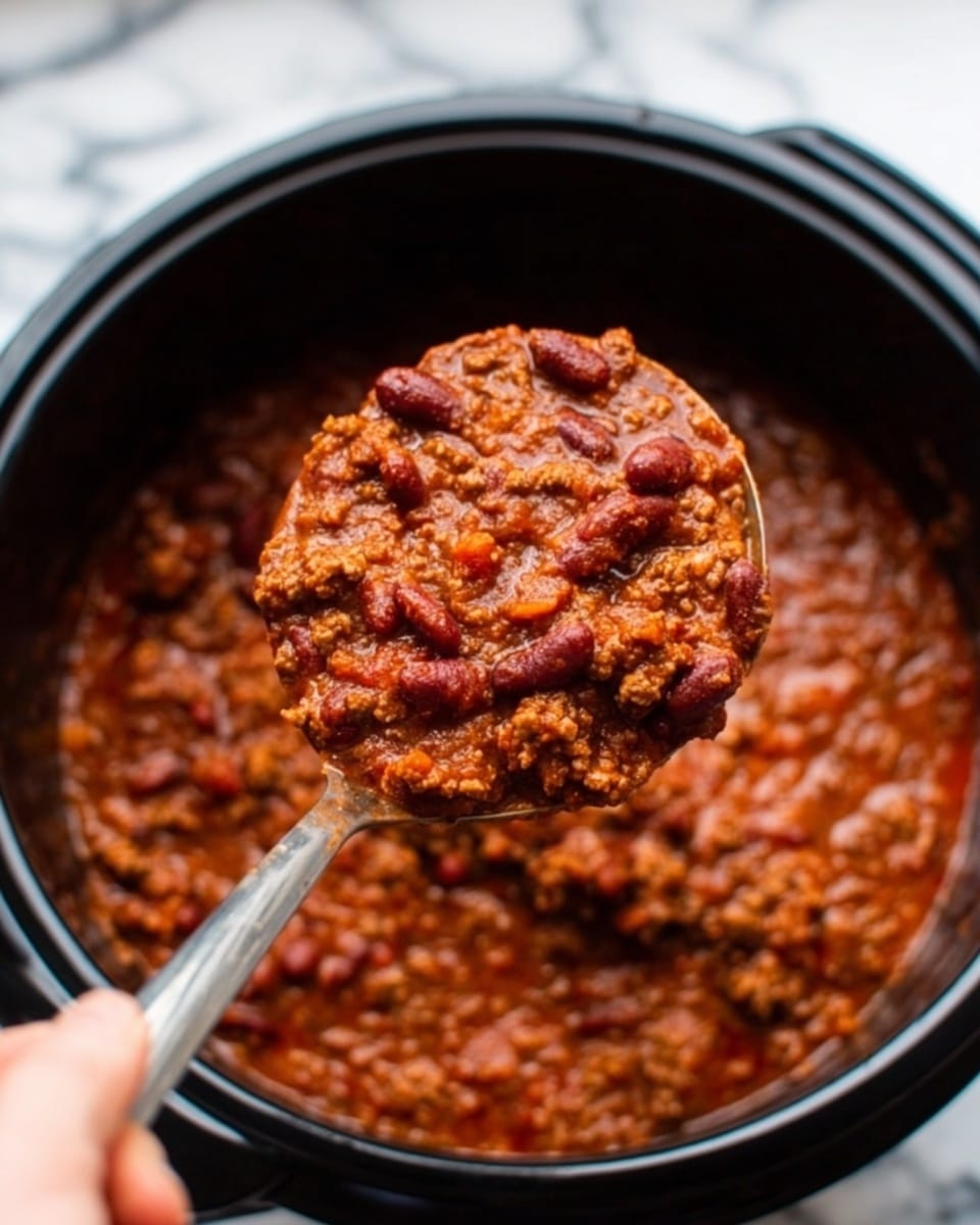 The image shows a black slow cooker filled with chili. The chili has a thick reddish-brown sauce layer with visible small chunks of cooked ground meat and kidney beans mixed in. Diced tomatoes and small pieces of green and orange vegetables are scattered throughout the mixture. A wooden spoon with a light natural color rests inside the cooker on the right side, partially submerged in the chili. The slow cooker sits on a white marbled surface. photo taken with an iphone --ar 4:5 --v 7