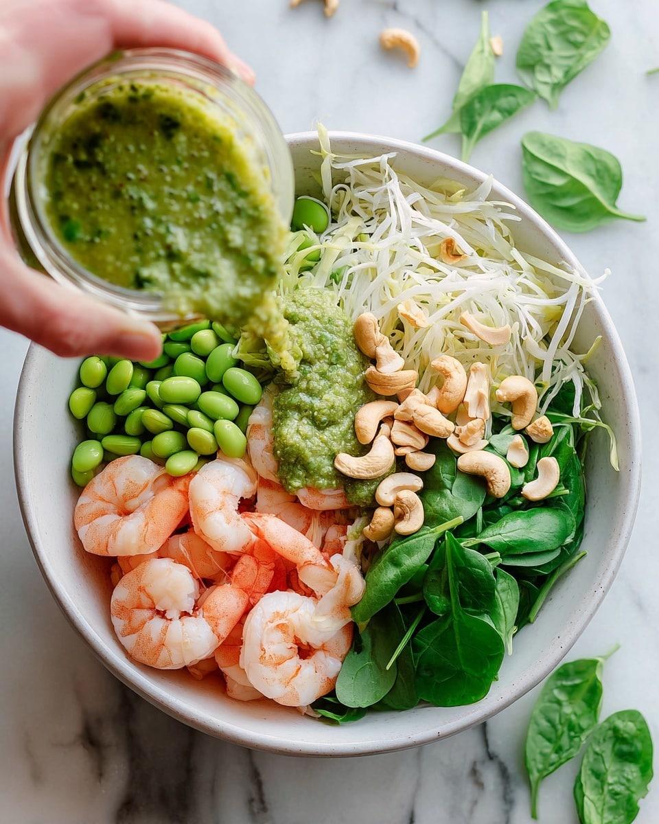 A white bowl holds a colorful shrimp salad placed on a white marbled surface. The bowl has four clear layers visible from top: bright green fresh spinach leaves on the right, lightly cooked pink shrimp on the bottom right, bright green edamame beans on the left, and thin white shredded cabbage at the bottom left. On top of the shrimp and cabbage are light brown cashew nuts, some whole and some broken. A thick green sauce with little herb bits is poured over the shrimp and cashews from a small clear glass jar held by a woman's hand coming in from the top left. Nearby on the surface are some loose spinach leaves, edamame beans, and shredded cabbage. photo taken with an iphone --ar 4:5 --v 7