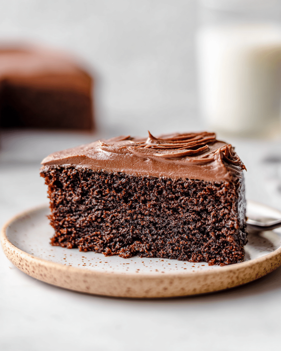 A single slice of chocolate cake is shown on a white plate with a slightly raised edge. The cake has two layers: a dense, moist dark brown base with a rough texture, and a thick layer of smooth, creamy chocolate frosting on top. The frosting is shiny and spread unevenly with visible soft ridges. The white marbled surface beneath the plate adds a clean and simple backdrop. In the blurred background, there is a glass with a white liquid, likely milk. Photo taken with an iphone --ar 4:5 --v 7