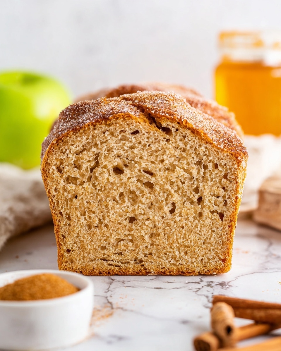A golden-brown loaf cake with a slightly cracked top is covered with a fine layer of sugar and cinnamon, giving it a rough texture. The cake has a thick, firm crust with a warm, baked color, sitting on parchment paper on a cooling rack. In the background, glass pitcher filled with amber liquid and two green apples are blurred, all set on a white marbled surface. photo taken with an iphone --ar 4:5 --v 7