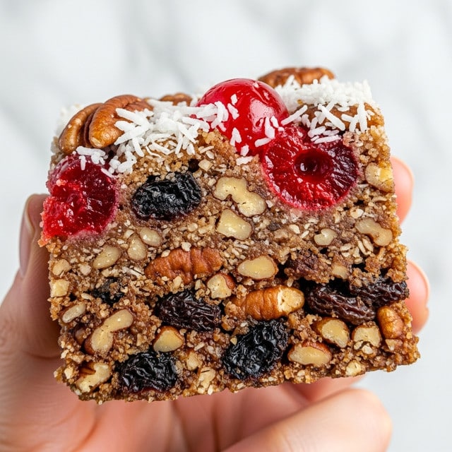 A close-up of a square granola bar showing multiple layers and textures: the base is a dense mix of brown pecan nuts and darker raisins, with bright red cherry pieces scattered on top, giving a pop of color. Small white coconut flakes add contrast and a rough texture on the surface. The granola bar is held by a woman's hand, and the background is a white marbled texture, making the colors stand out clearly. photo taken with an iphone --ar 4:5 --v 7