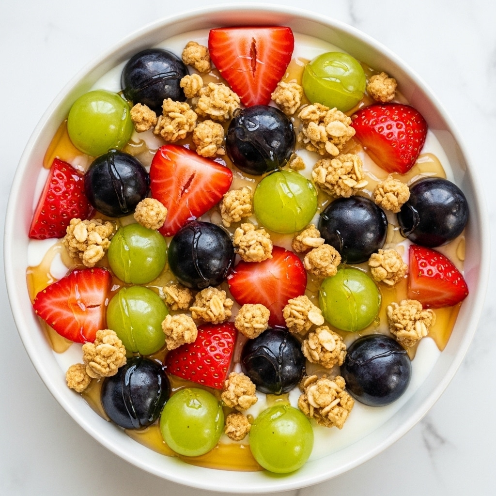 The dish is a layered dessert in a clear square glass bowl set on a white marbled surface. The bottom layer is a crumbly light brown base, possibly crushed cookies or graham crackers. Above that is a thick, fluffy white cream layer with a smooth texture. The top layer is generously decorated with fresh whole green grapes and red grapes, sliced strawberries showing their bright red interior, small scattered nut pieces, and bits of crumbled light brown topping. A glossy caramel sauce is drizzled unevenly over the top, adding a shiny golden-brown color that contrasts with the fresh fruit and cream. photo taken with an iphone --ar 4:5 --v 7