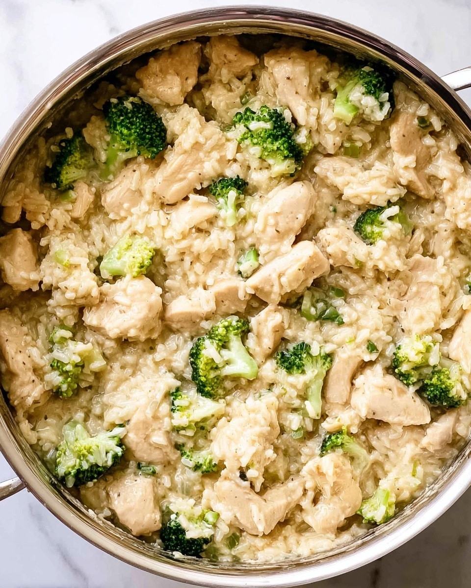The image shows a close-up view of a creamy chicken and broccoli rice dish in a silver pot. It has three main layers: tender light beige chicken pieces that look moist, small cuts of bright green broccoli florets scattered evenly, and soft, creamy beige rice mixed throughout. The rice has a smooth texture surrounding the chicken and broccoli. The silver pot occupies the right and bottom edges with some sauce residue on its sides, while the dish sits on a white marbled surface. photo taken with an iphone --ar 4:5 --v 7