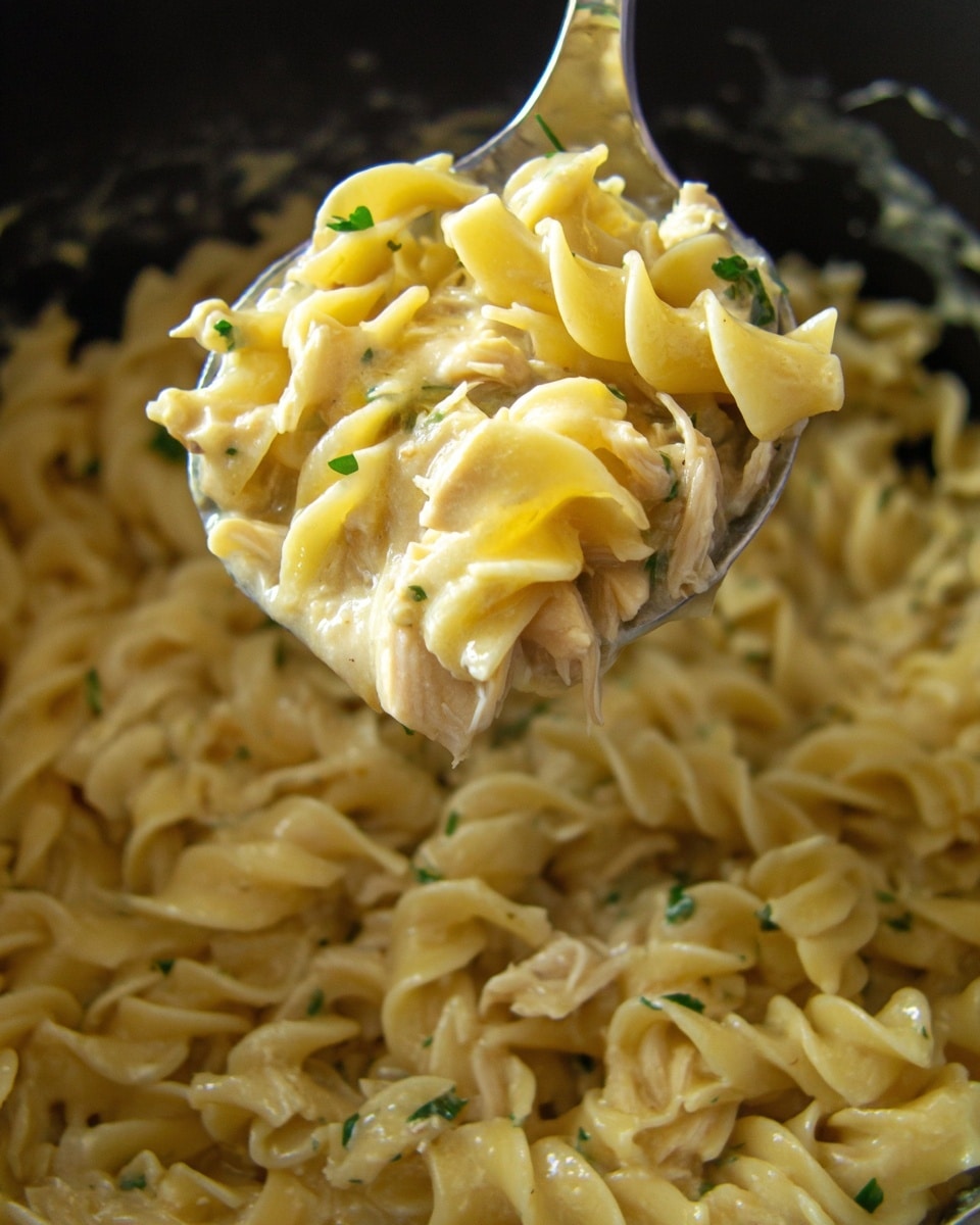 A close-up view of a shiny metal spoon lifting a creamy mixture of egg noodles and bits of chicken from a dark pot. The noodles are soft with a smooth pale yellow color mixed with slightly darker creamy sauce, giving a glossy and moist texture. Small green herb pieces are scattered lightly across the noodles, adding a touch of color. The noodles have a twisted, curly shape and fill the pot in the background, which is out of focus. The spoon is centered in the image, emphasizing the thick and rich noodle blend. Photo taken with an iphone --ar 4:5 --v 7