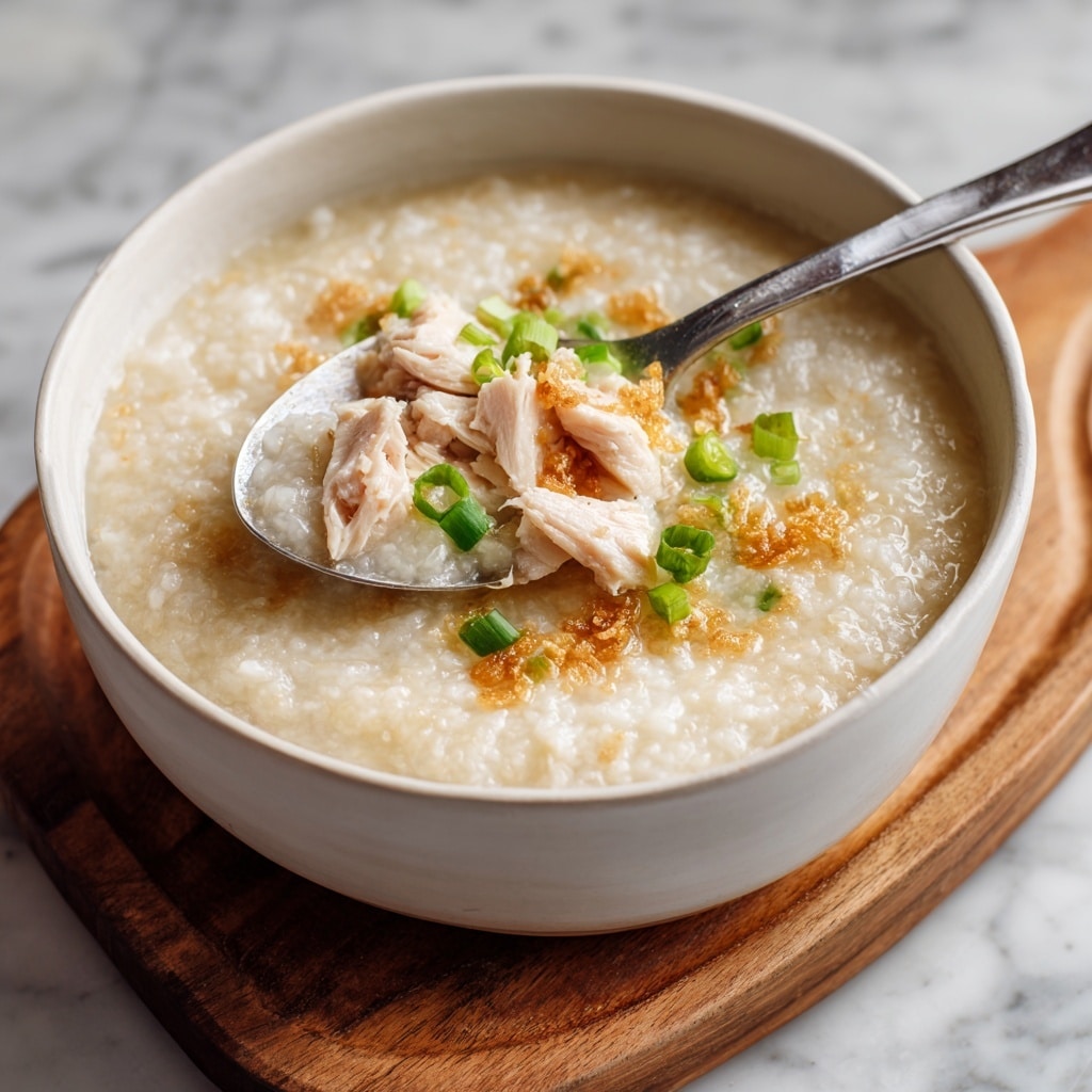 A white ceramic bowl holds a warm, thick rice porridge with a pale beige color and a soft, slightly lumpy texture. On top, there are small pieces of tender light pink chicken placed slightly to the right side of the bowl, partially lifted by a shiny silver spoon. Scattered green onion slices add a bright green contrast near the center. Tiny crispy golden-brown bits are sprinkled around the chicken, adding a crunchy element. The bowl sits on a wooden board, and the scene is set against a white marbled surface. Photo taken with an iphone --ar 4:5 --v 7