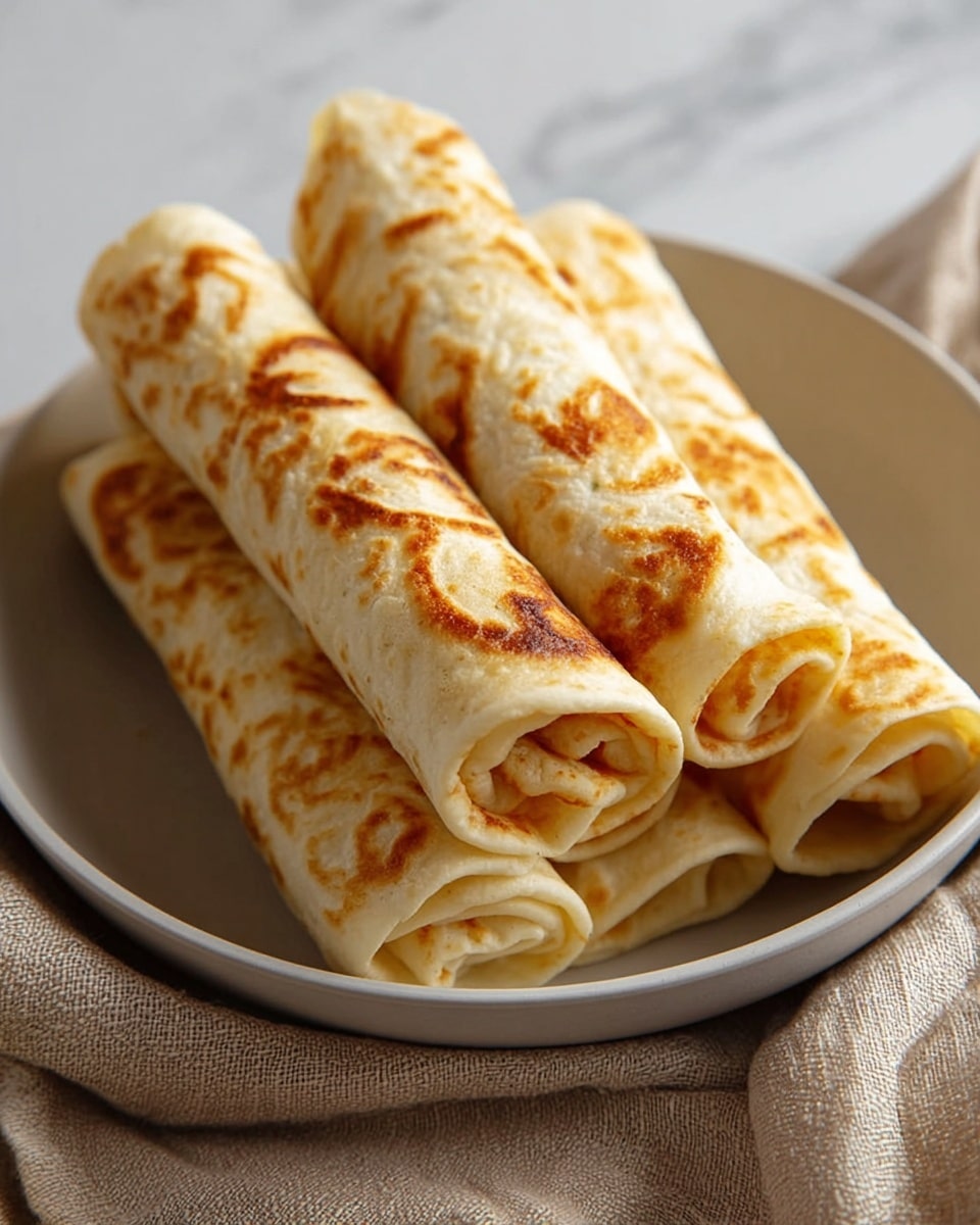 The image shows five rolled flatbreads stacked together in a round white bowl. Each flatbread is light beige with golden brown spots from cooking, showing a thin, soft texture with slight layering on the edges. The rolls are placed closely, some overlapping each other, with the rolled ends facing outward. The bowl sits on a neutral-colored cloth on a white marbled surface in soft natural light. photo taken with an iphone --ar 4:5 --v 7