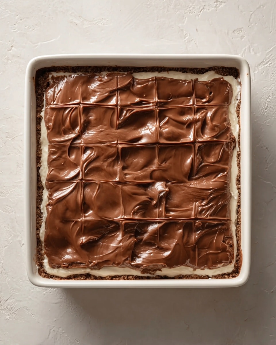 A square white baking dish holds a layered dessert with three visible layers. The bottom layer is a crumbly brown crust, slightly visible at the edges. Above that is a smooth, creamy white layer filling most of the dish. The top layer is thick, glossy milk chocolate, spread evenly but with natural swirls and some textured lines forming a grid-like pattern across the surface. The dish is set on a surface with a white marbled texture. photo taken with an iphone --ar 4:5 --v 7