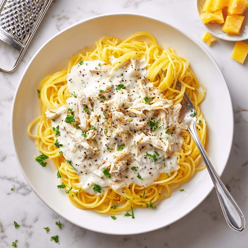 A white plate filled with a base layer of thin, yellow spaghetti noodles, loosely piled in a circular shape. On top, there is a creamy white sauce with visible black pepper specks and shredded chicken pieces coated in the sauce, scattered evenly over the noodles. Small green parsley leaves are sprinkled across the dish, adding a touch of color. A silver fork is resting on the right side of the plate, slightly twirling some noodles. The plate sits on a white marbled surface, with a metal grater and small yellow cheese chunks in the background. photo taken with an iphone --ar 4:5 --v 7