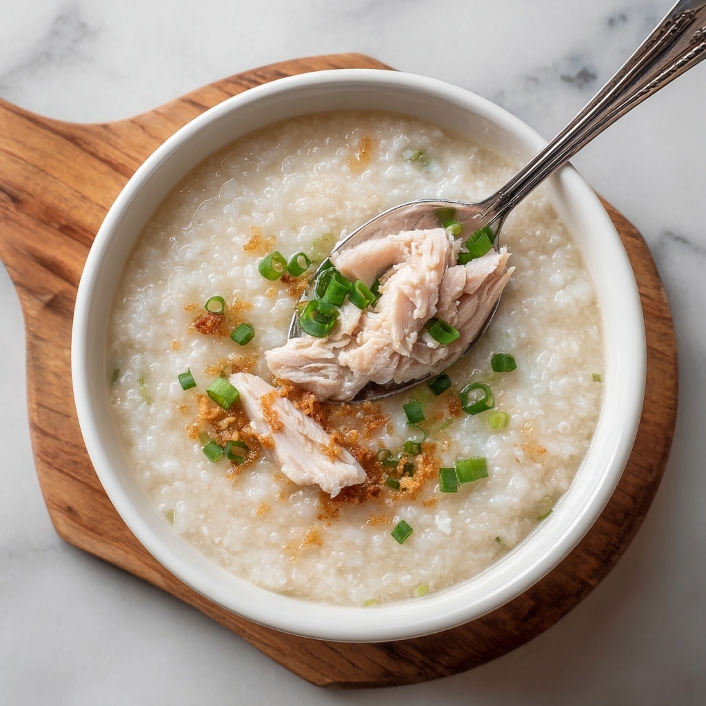 The image shows a white bowl filled with three main layers: a thick, creamy rice porridge in a light beige color forms the base, with a slightly glossy and soft texture; on top of this, there are small green rings of fresh sliced scallions scattered on one side; the third layer features tender cooked chicken pieces in a light pinkish-beige color partially submerged in the porridge near a spoon lifting some chicken; the bowl is placed on a wooden board, all set against a white marbled texture surface. photo taken with an iphone --ar 4:5 --v 7