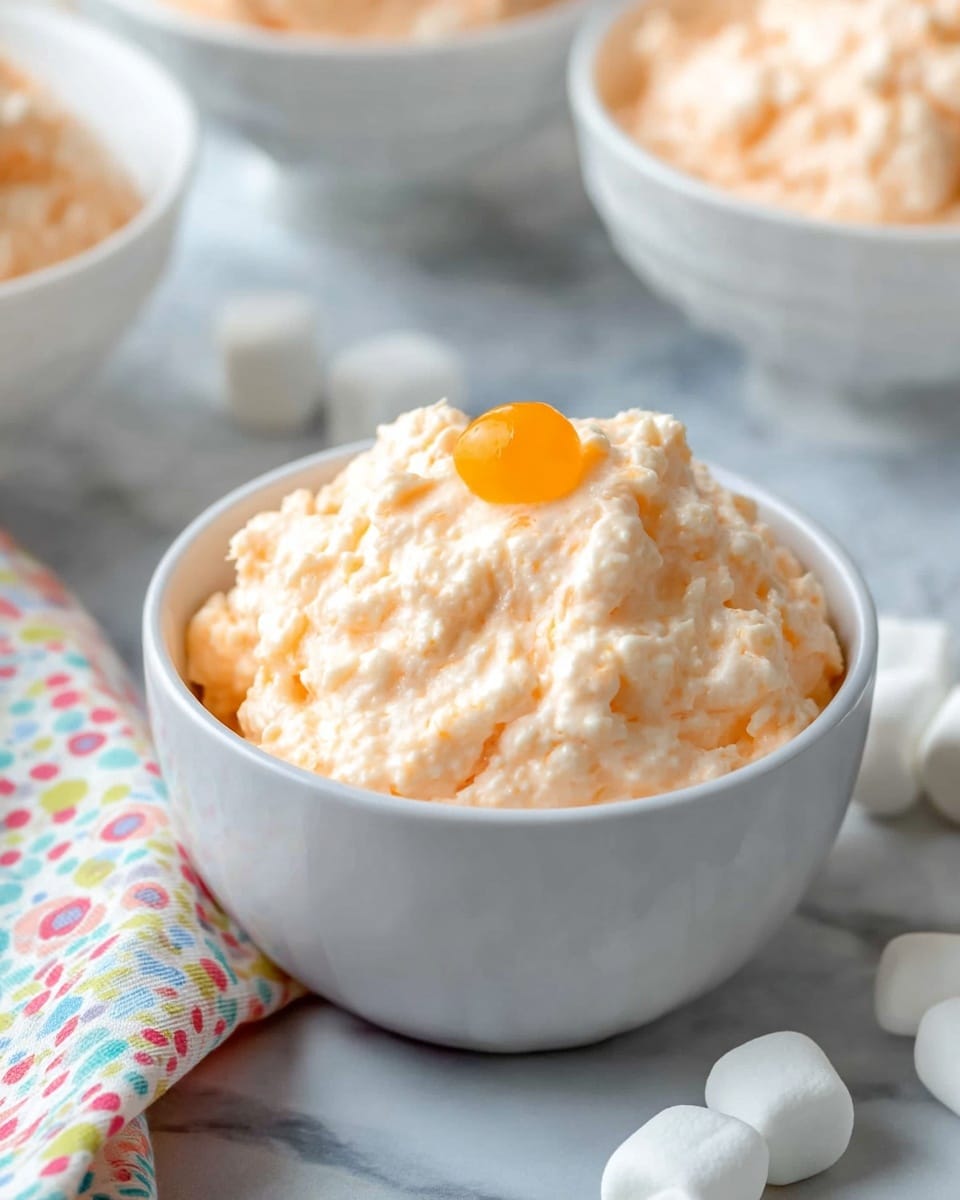 A white bowl filled with a creamy, light orange mixture that has a chunky texture, showing small lumps throughout. Mixed inside are several bright orange mandarin segments scattered around and partially submerged in the creamy layer. The bowl sits on a white marbled surface with a colorful cloth nearby. The creamy layer forms the whole dish, with its soft and thick texture making the mandarin slices stand out with their glossy smoothness. Photo taken with an iphone --ar 4:5 --v 7