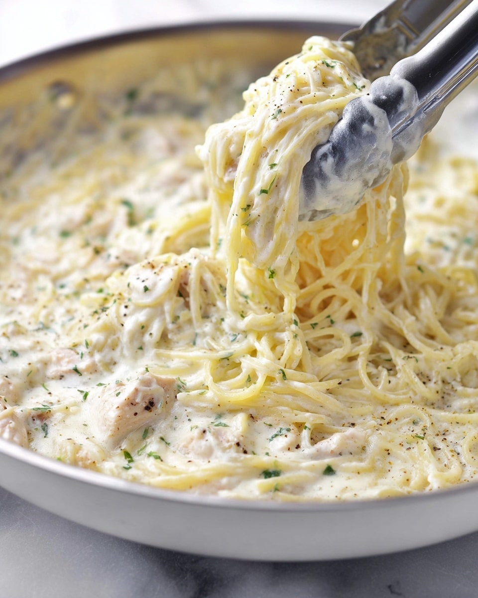 The image shows a close-up of creamy white pasta being lifted by silver tongs, revealing soft spaghetti coated in a thick, cheesy white sauce mixed with small pieces of chicken and specks of green herbs. The sauce looks smooth and rich, clinging to the noodles, with hints of black pepper sprinkled on top. The pasta is in a white pan resting on a white marbled surface, giving a fresh clean look. photo taken with an iphone --ar 4:5 --v 7