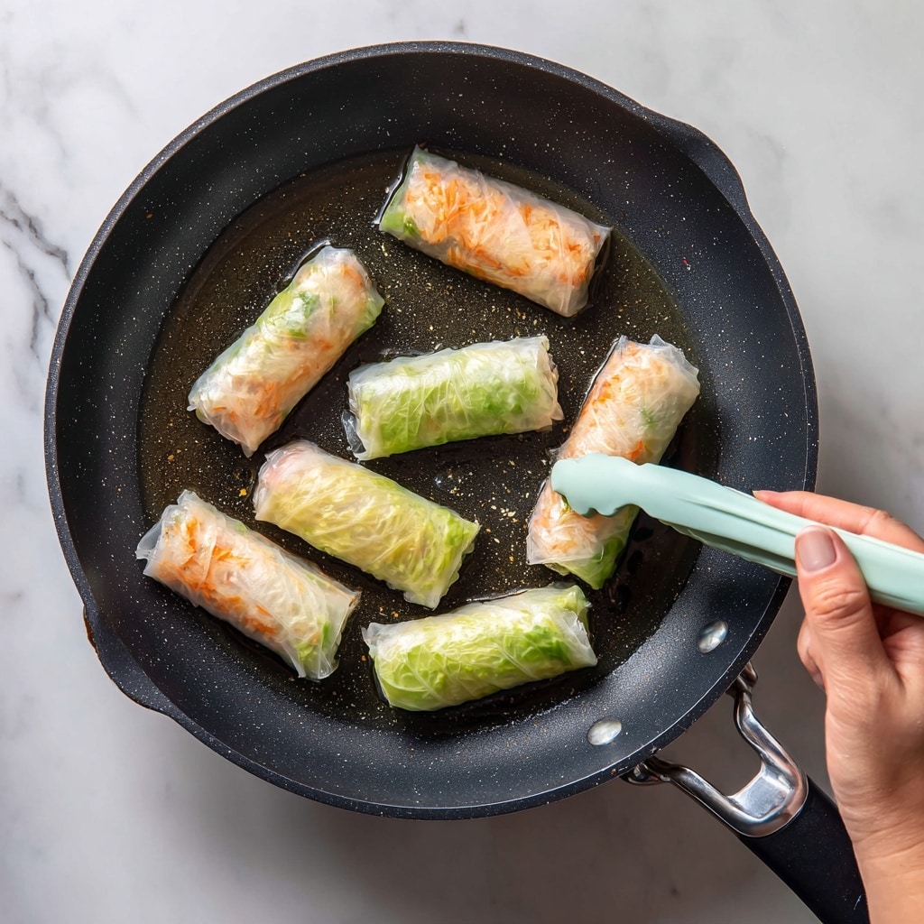 In a dark non-stick pan, there are nine rolled translucent rice paper rolls cooking lightly in oil. Each roll shows a mix of light orange and white colors inside, likely from a filling, with one roll wrapped in a pale green leafy layer visible underneath the rice paper. A pair of light green tongs held by a woman’s hand is lifting one of the rolls. The rice paper has a shiny, slightly wet texture and the pan surface is dotted with small oil drops. The scene sits on a white marbled surface. photo taken with an iphone --ar 4:5 --v 7
