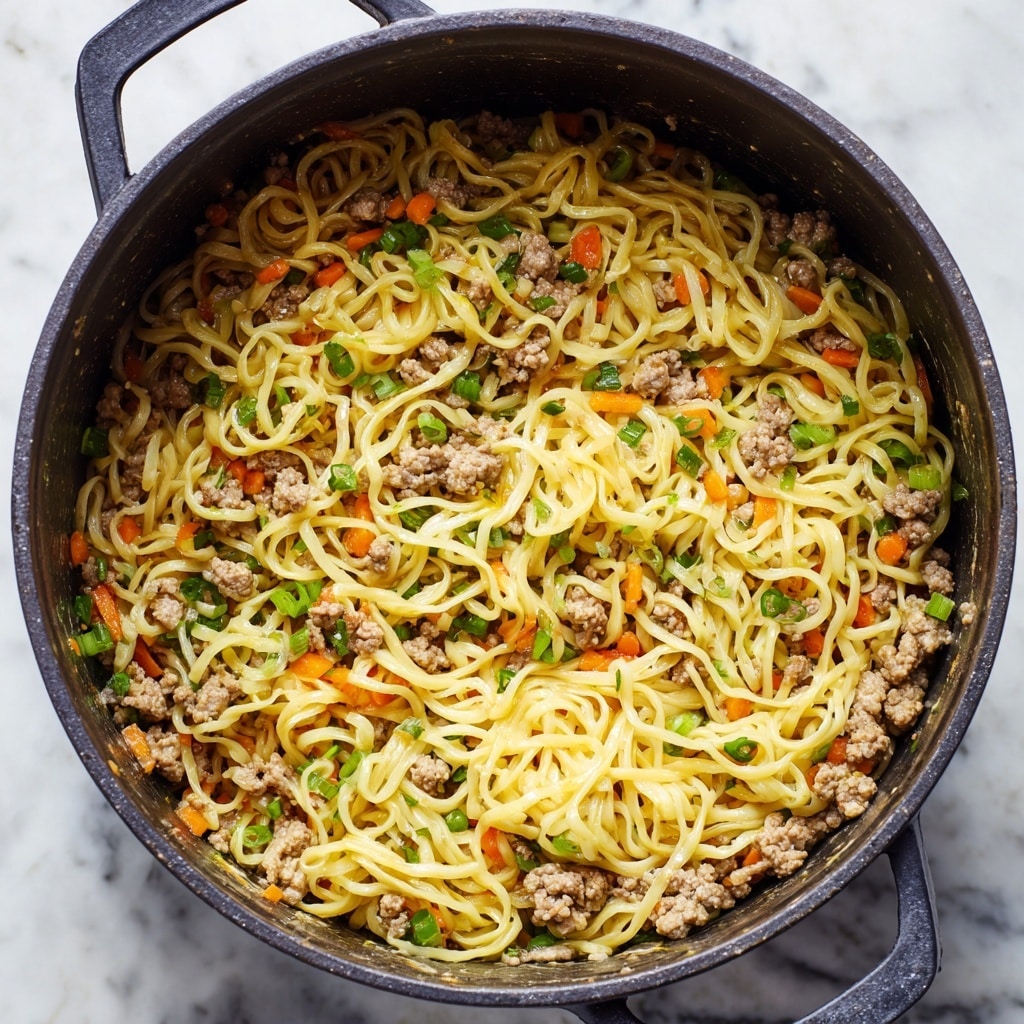 A close-up view inside a dark gray cooking pot shows cooked yellow noodles mixed well with small crumbled pieces of light brown ground meat, scattered bright green onion pieces, and small orange carrot chunks. The noodles have a smooth and slightly shiny texture, coated lightly with sauce, while the vegetables and meat create a varied texture throughout. The depth of the pot holds the mixture together, and the background has a white marbled texture. photo taken with an iphone --ar 4:5 --v 7